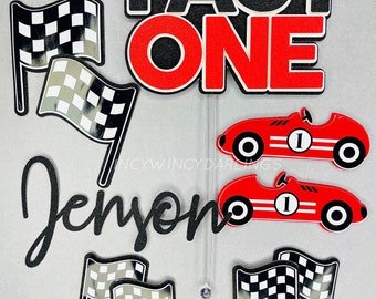 May include: A cake topper set with a racing theme. The words "FAST ONE" in black and red are at the top. Below are two red race cars with the number "1" on them, checkered flags, and the name "Jenson".