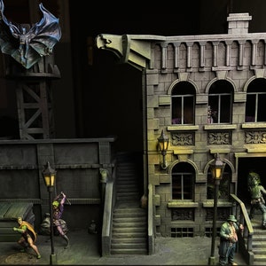 Könnte beinhalten: Eine Miniatur-Stadtlandschaft mit einem dunkelgrauen Gebäude, einem grauen Gebäude mit einer Gargoyle und einem hohen Bauwerk, das mit einer Fledermausfigur gekrönt ist. Die Gebäude haben Fenster und Türen, und es stehen kleine Figuren von Menschen auf dem Boden.