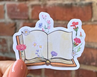 May include: An open book sticker with a white background and a brown cover. The book is decorated with colorful flowers, including pink, red, purple, and blue.