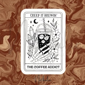 以下が含まれることがあります： 透明なカップに入ったコーヒーを、骸骨の手が持っている白黒のステッカー。「CREEP IT BREWIN'」と「THE COFFEE ADDICT」の文字。クリスタル、花、三日月などの装飾が施されています。