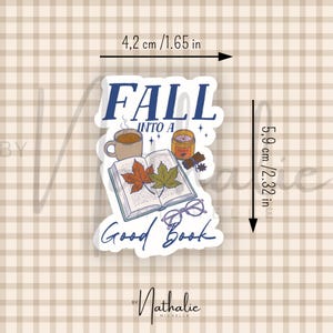 May include: A die-cut sticker featuring an open book with two autumn leaves, a mug, a candle, and glasses. The text reads FALL INTO A GOOD BOOK. It measures 1.65 inches wide by 2.32 inches high, as indicated by the visible measurement markings.