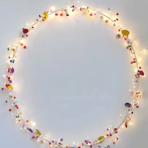Puede incluir: Un círculo de alambre blanco con una cadena de luces blancas de hadas envuelta alrededor. Las luces están decoradas con cuentas de colores, lentejuelas y otros pequeños artículos decorativos.