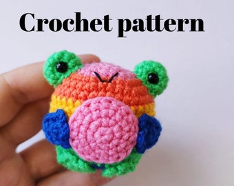 May include: A crocheted rainbow frog holding a pink ball. The frog has green eyes and a pink nose. The frog is a small amigurumi toy.