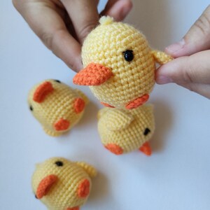 May include: A collection of handmade crochet yellow chicks with orange beaks and feet. The chicks have black safety eyes and small wings. The chicks are displayed on a white surface.