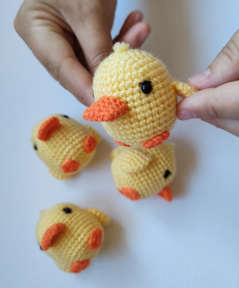 May include: A collection of handmade crochet yellow chicks with orange beaks and feet. The chicks have black safety eyes and small wings. The chicks are displayed on a white surface.