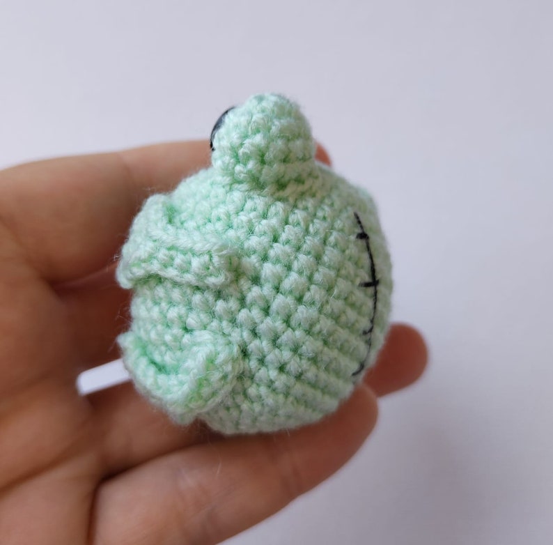 May include: A small, crocheted green frog with black eyes. The frog is sitting on a hand.