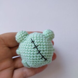 May include: A small, green crocheted stuffed animal with a stitched-up seam across the back. The animal has two small, round ears.