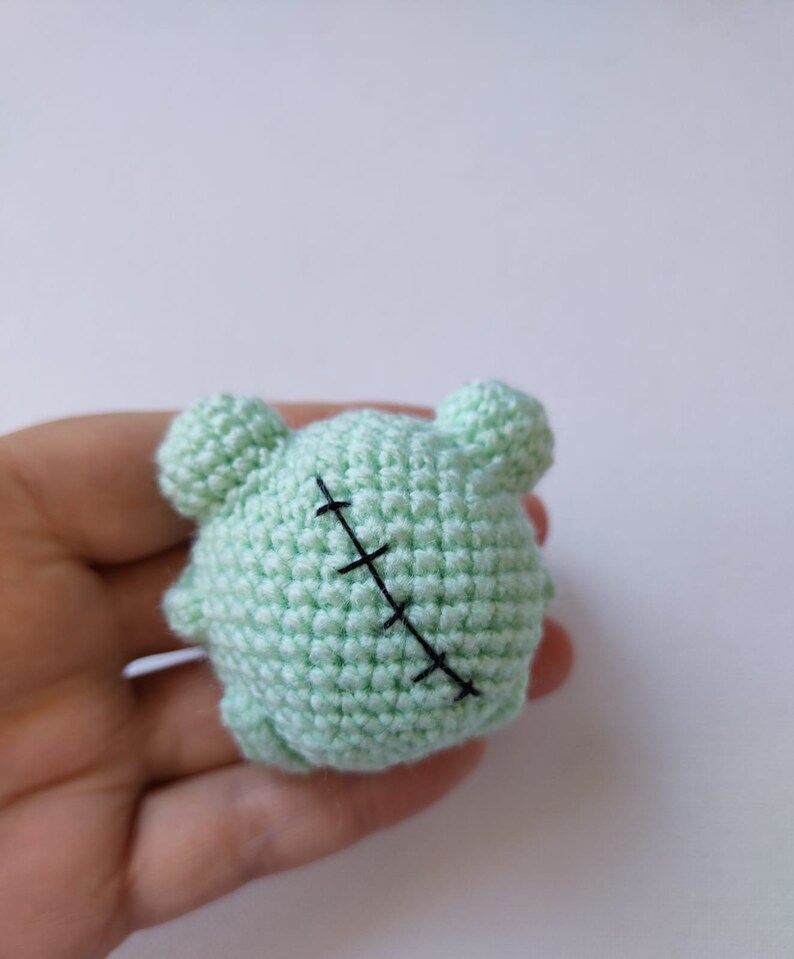 May include: A small, green crocheted stuffed animal with a stitched-up seam across the back. The animal has two small, round ears.