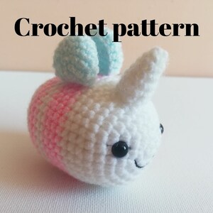 May include: Crochet pattern for a white and pink amigurumi bee with blue wings. The bee has black eyes and a smiling mouth.