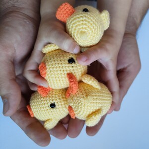 May include: Three small, crocheted yellow ducklings with orange beaks and feet, and black eyes. The ducklings are held in a person's hands, with a child's hands also visible. The ducklings are a handmade craft item.