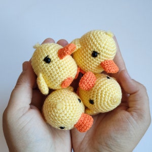 May include: Four handmade crochet ducklings in a person's hands. The ducklings are yellow with orange beaks and feet, and black eyes. The crochet work is detailed, and the ducklings appear soft and cuddly.