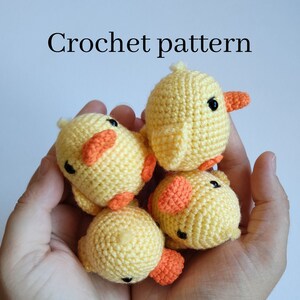 Op de afbeelding: Vier kleine, gele gehaakte eendjes met oranje snavels en voetjes worden in de handen van een persoon gehouden. De tekst "Crochet pattern" staat boven de eendjes. De eendjes hebben zwarte ogen en kleine vleugels.