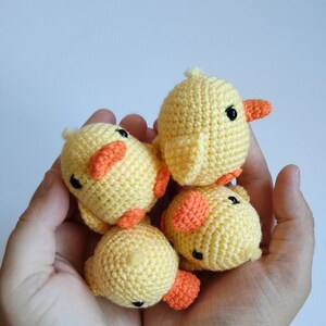 May include: Four small, handmade crochet ducklings. Each duckling is yellow with orange beaks and feet, and black eyes. The ducklings are held in a cupped hand against a plain white background. The crochet work is detailed and the ducklings appear soft.