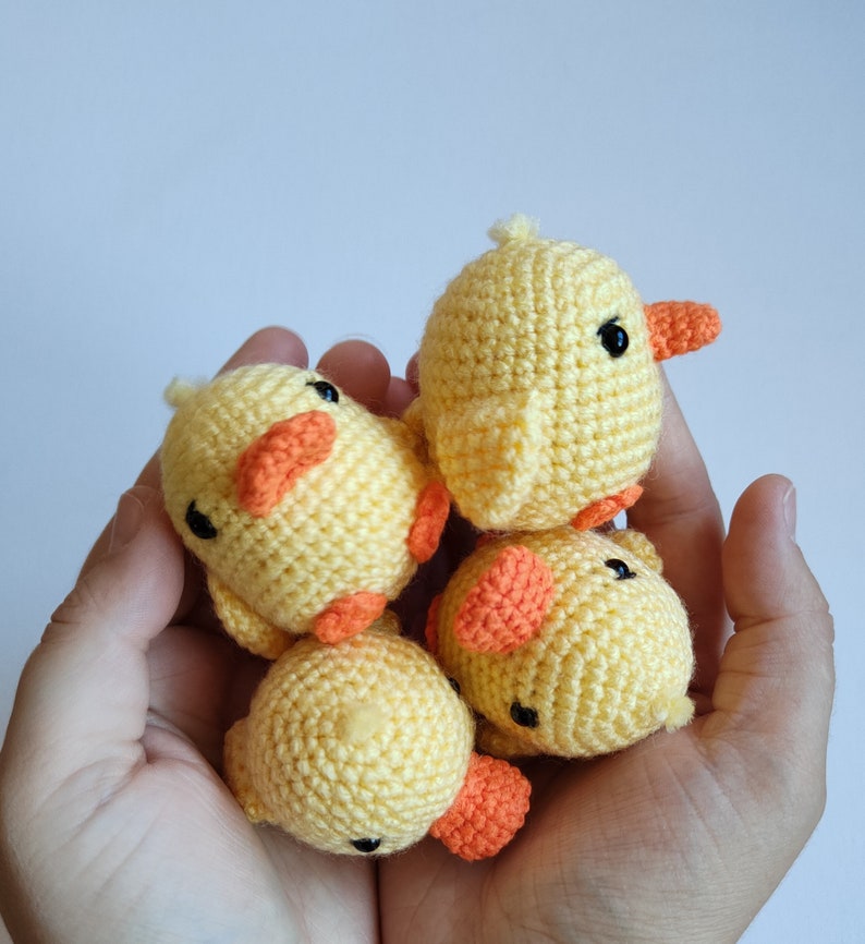 May include: Four small, handmade crochet ducklings. Each duckling is yellow with orange beaks and feet, and black eyes. The ducklings are held in a cupped hand against a plain white background. The crochet work is detailed and the ducklings appear soft.