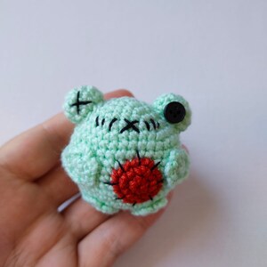 May include: A small crocheted green frog with black eyes and a red heart on its belly.