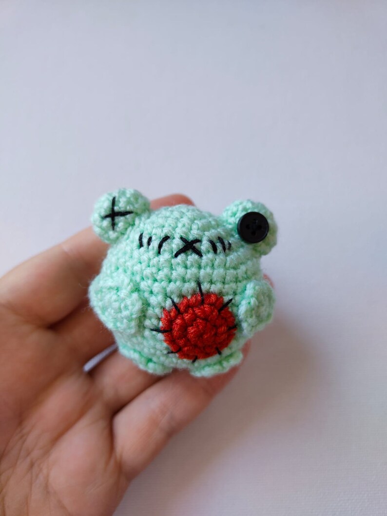 May include: A small crocheted green frog with black eyes and a red heart on its belly.
