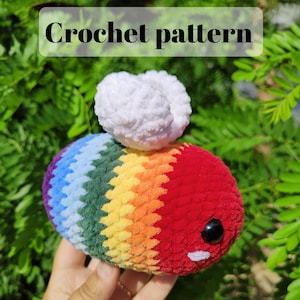 Puede incluir: Una abeja de crochet arcoíris con una nube blanca de crochet en la espalda. La abeja tiene un ojo negro y una pequeña boca blanca.