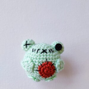 May include: A small, green crocheted frog with a red heart on its chest and a black button for an eye.