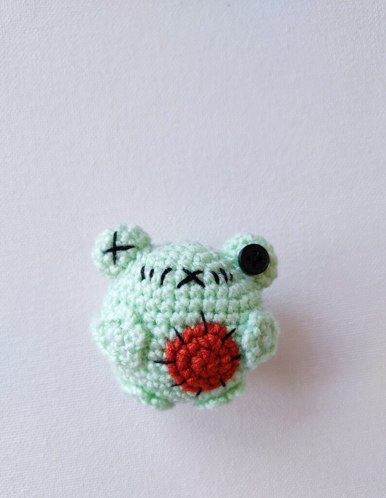 May include: A small, green crocheted frog with a red heart on its chest and a black button for an eye.