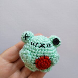 May include: A small green crocheted frog with a red heart on its belly and a black button for an eye.