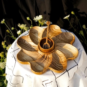 May include: A wooden serving platter with a flower-like design, featuring a central bowl and a honey dipper. The platter is made of light-colored wood with visible grain patterns. The honey dipper is placed inside the bowl.