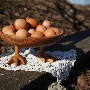 May include: A wooden bowl filled with brown eggs, resting on a white lace doily. The bowl is supported by three carved wooden chicken feet. The eggs are a light brown color, and the bowl has a natural wood grain.