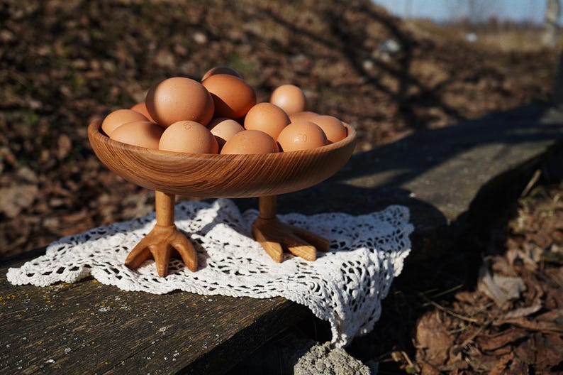 May include: A wooden bowl filled with brown eggs, resting on a white lace doily. The bowl is supported by three carved wooden chicken feet. The eggs are a light brown color, and the bowl has a natural wood grain.