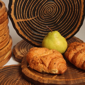May include: A set of wooden serving trays featuring a tree ring design. The trays are crafted from dark wood with a natural finish. The trays are stacked, with a pear and two croissants on the top tray.
