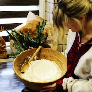 Peut inclure: Une femme mélange de la farine dans un grand bol en bois avec une cuillère en bois. Le bol est rempli de farine blanche et la femme tient le bol avec les deux mains.