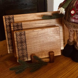 May include: Three wooden cutting boards with a decorative design on the edges. The boards are stacked on top of each other and are sitting on a wooden surface. A small green plant is in front of the boards.
