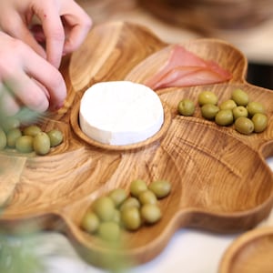 May include: A wooden, leaf-shaped serving platter filled with olives, cheese, and meat. The platter has multiple compartments and a natural wood grain finish. The image shows a person's hands arranging the food on the platter.