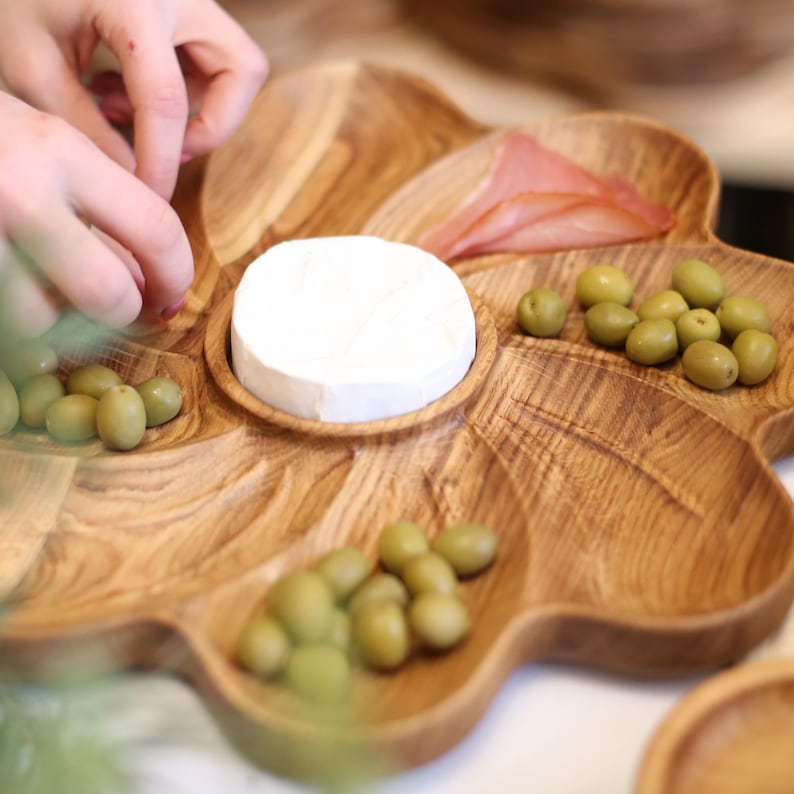 May include: A wooden, leaf-shaped serving platter filled with olives, cheese, and meat. The platter has multiple compartments and a natural wood grain finish. The image shows a person's hands arranging the food on the platter.