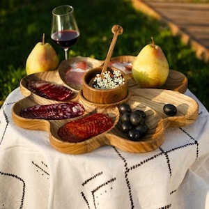 May include: A wooden serving platter with a flower-like design, filled with sliced meats, olives, and a small bowl of flowers. Two pears and a glass of red wine are also present. The platter is on a white tablecloth.