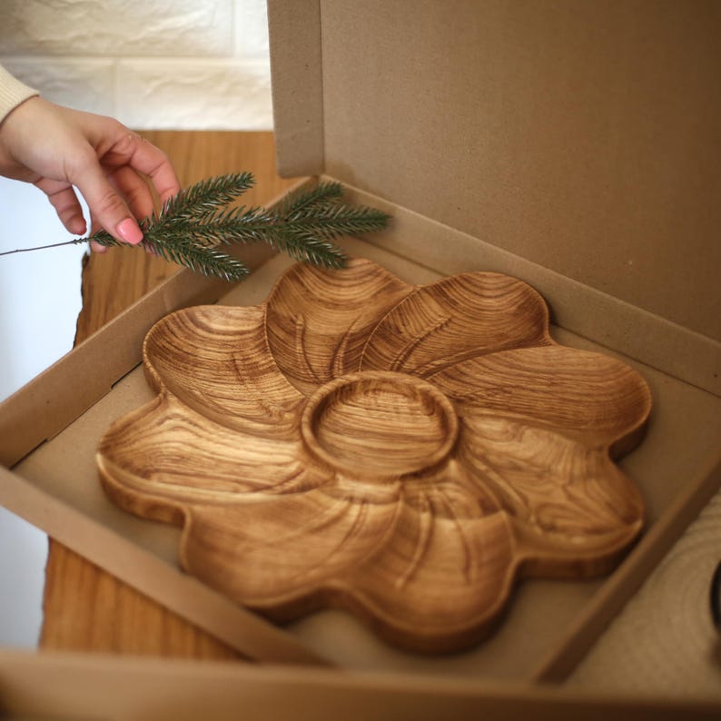 May include: A wooden serving platter in the shape of a flower, with a central dip bowl and petal-shaped sections. The platter is inside a cardboard box. The wood grain is visible, and the color is a light brown.