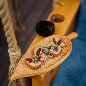 May include: A wooden serving tray shaped like a leaf with four pieces of bread topped with pate, sun-dried tomatoes, and herbs. The tray is on a wooden surface with a glass of red wine in the background.
