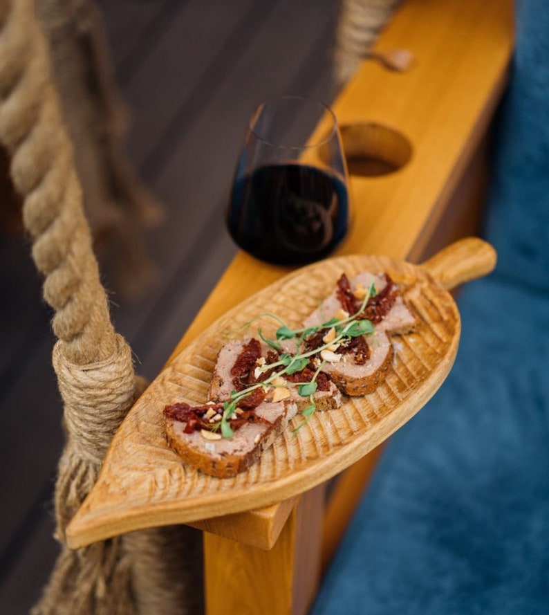 May include: A wooden serving tray shaped like a leaf with four pieces of bread topped with pate, sun-dried tomatoes, and herbs. The tray is on a wooden surface with a glass of red wine in the background.