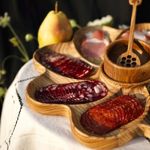 Peut inclure: Un plateau de service en bois avec plusieurs compartiments, présentant une variété de viandes tranchées, dont du salami et du prosciutto. Le plateau comprend également un petit bol en bois avec un cuillère à miel.