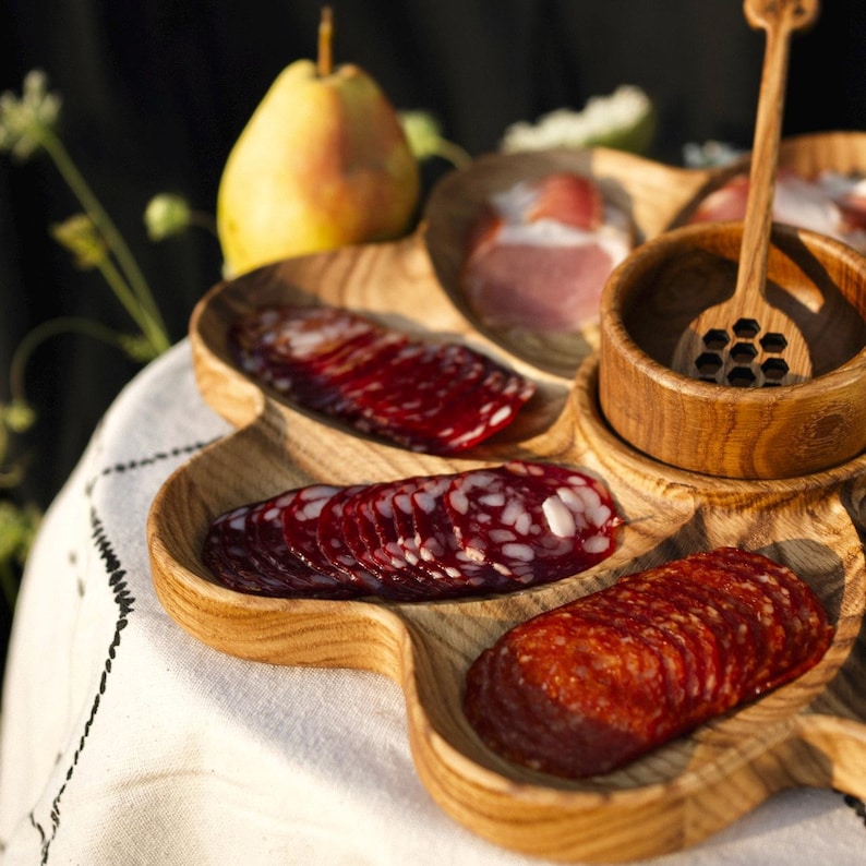 May include: A wooden serving platter with multiple compartments, filled with sliced meats, a small wooden bowl, and a honey dipper. A yellow pear is in the background. The platter is on a white tablecloth.