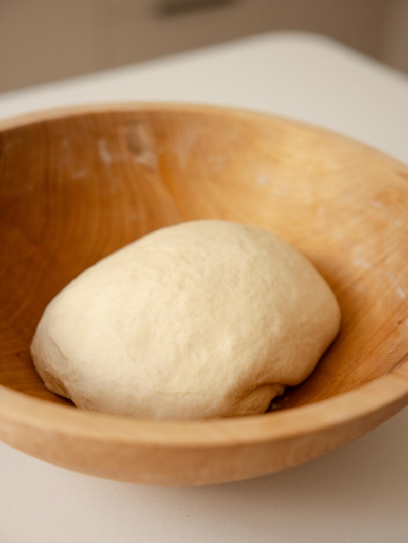 K&ouml;nnte beinhalten: Ein glatter, runder Teigballen in hellem Beige, der in einer gro&szlig;en, unbehandelten hellbraunen Holzsch&uuml;ssel liegt. Der Teig hat eine weiche, leicht strukturierte Oberfl&auml;che und die Sch&uuml;ssel zeigt durchgehend sichtbare Holzmaserungen.
