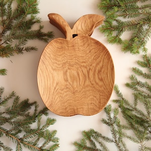Puede incluir: Un plato de servir de madera con forma de manzana hecho de madera de roble. El plato es de color marrón claro y tiene un patrón de veta de madera natural. El plato es perfecto para servir aperitivos, salsas u otros artículos pequeños.