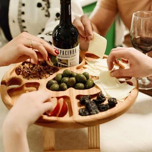 Op de afbeelding: Een houten serveerschaal met meerdere compartimenten, met een fles rode wijn, olijven, kaas, noten en gedroogd fruit. De schaal staat op een kleine houten standaard.