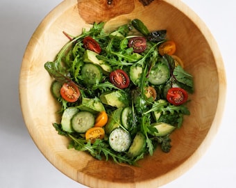 Könnte beinhalten: Eine große, runde, hellbraune Holzschüssel gefüllt mit einem frischen Salat. Der Salat enthält grüne Rucolablätter, Gurkenscheiben, grüne Avocadostücke und halbierte Kirschtomaten in den Farben Rot, Orange und Dunkellila.