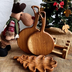 May include: Wooden serving boards and a leaf-shaped tray are displayed. Two round boards with handles are stacked, and a leaf-shaped tray sits in front. A wooden rack and a Christmas tree are in the background. The items are made of light brown wood.