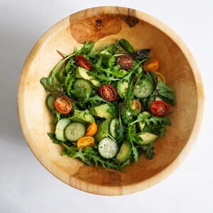 K&ouml;nnte beinhalten: Eine gro&szlig;e, runde, hellbraune Holzsch&uuml;ssel gef&uuml;llt mit einem frischen Salat. Der Salat enth&auml;lt gr&uuml;ne Rucolabl&auml;tter, Gurkenscheiben, gr&uuml;ne Avocadost&uuml;cke und halbierte Kirschtomaten in den Farben Rot, Orange und Dunkellila.