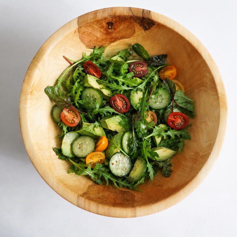 K&ouml;nnte beinhalten: Eine gro&szlig;e, runde, hellbraune Holzsch&uuml;ssel gef&uuml;llt mit einem frischen Salat. Der Salat enth&auml;lt gr&uuml;ne Rucolabl&auml;tter, Gurkenscheiben, gr&uuml;ne Avocadost&uuml;cke und halbierte Kirschtomaten in den Farben Rot, Orange und Dunkellila.