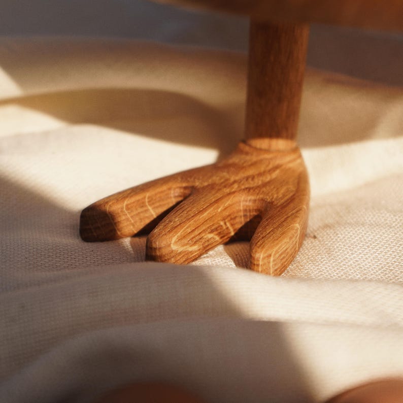 May include: Close-up of a wooden serving stand base. The base is crafted from light brown wood, featuring a hand-shaped design with three fingers. The wood grain is visible, and the stand is set against a soft, off-white fabric background.