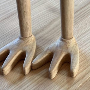 May include: Two wooden salad servers with a fork-like design. The handles are cylindrical, and the fork ends have three tines. The wood has a natural, light brown color, and the servers are resting on a wooden surface.