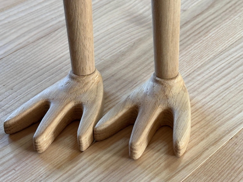 May include: Two wooden salad servers with a fork-like design. The handles are cylindrical, and the fork ends have three tines. The wood has a natural, light brown color, and the servers are resting on a wooden surface.