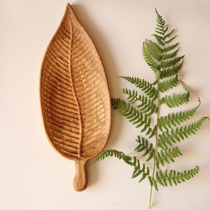 May include: A wooden leaf-shaped serving tray with a carved design. The tray is made of light brown wood and has a natural finish. The tray is sitting on a white surface.
