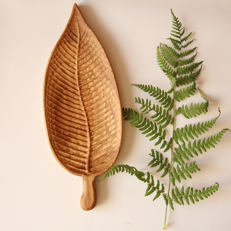May include: A wooden leaf-shaped serving tray with a carved design. The tray is made of light brown wood and has a natural finish. The tray is sitting on a white surface.